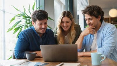 Drei junge Erwachsene betrachten lächelnd den Bildschirm eines Laptops am Tisch.