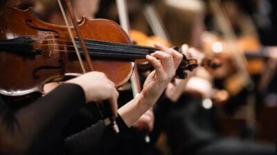 Nahaufnahme der Hände einer Geigerin beim Spielen der Violine mit Orchestermusikern im Hintergrund