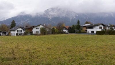 Grüne Wiese vor mehreren modernen Wohnhäusern mit wolkenverhangenem Bergmassiv im Hintergrund.