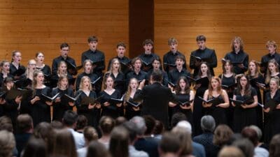 Jugendchor in schwarzen Kleidern singt mit Notenheften auf Bühne vor Publikum