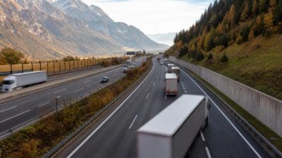 Mehrere Lastwagen fahren auf einer mehrspurigen Autobahn durch ein herbstliches Tal mit Bergen und schneebedeckten Gipfeln