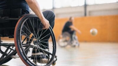 Person im Sportrollstuhl greift an das Rad, im Hintergrund ein weiterer Rollstuhlfahrer und Ball in der Halle
