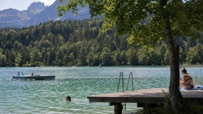 Eine Frau sitzt auf einem Holzsteg am See mit Schwimmern und Booten vor Bergen und Wald