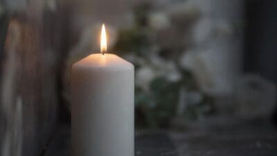 Brennende weiße Kerze im Vordergrund, unscharfer Blumenstrauß im Hintergrund