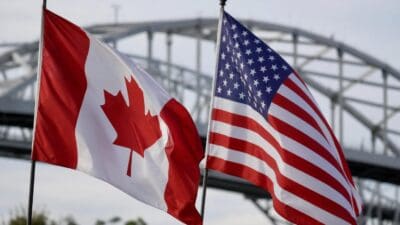 Kanadische und US-amerikanische Flagge wehen vor einer Stahlbrücke im Hintergrund