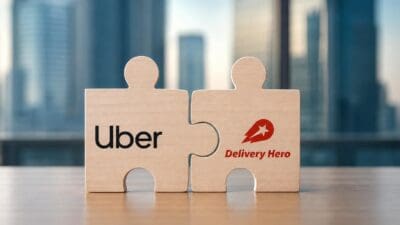 Zwei Holzpuzzle-Teile mit Uber- und Delivery-Hero-Logo stehen zusammen auf einem Tisch