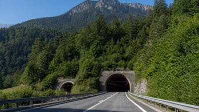 Asphaltstraße mit Leitplanken führt in einen Tunnel in bewaldeter Berglandschaft unter blauem Himmel.