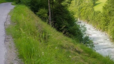 Asphaltierter Weg verläuft an steilem Grashang oberhalb eines reißenden Flusses in grüner Landschaft.
