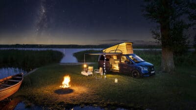 Dunkelblauer Mercedes-Benz Marco Polo Campervan mit beleuchtetem Aufstelldach steht am See bei einem Lagerfeuer