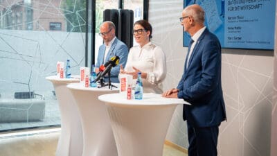 Eine Frau in weißer Bluse spricht gestikulierend an Stehtischen mit Mikrofonen, zwei Männer in Anzügen hören zu.