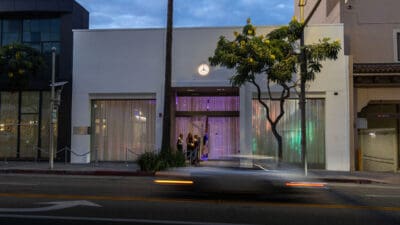 Moderne weiße Fassade eines Gebäudes mit beleuchtetem Mercedes-Logo, Personen am Eingang und einem vorbeifahrenden Auto