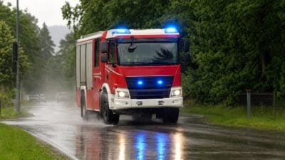 Rotes Feuerwehrauto der Feuerwehr mit blauem Blaulicht und Scheinwerfern auf einer nassen Straße im Regen