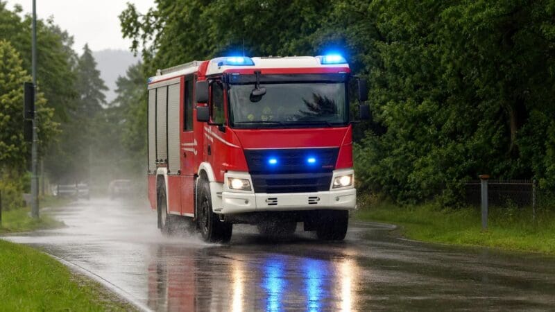 Rotes Feuerwehrauto der Feuerwehr mit blauem Blaulicht und Scheinwerfern auf einer nassen Straße im Regen
