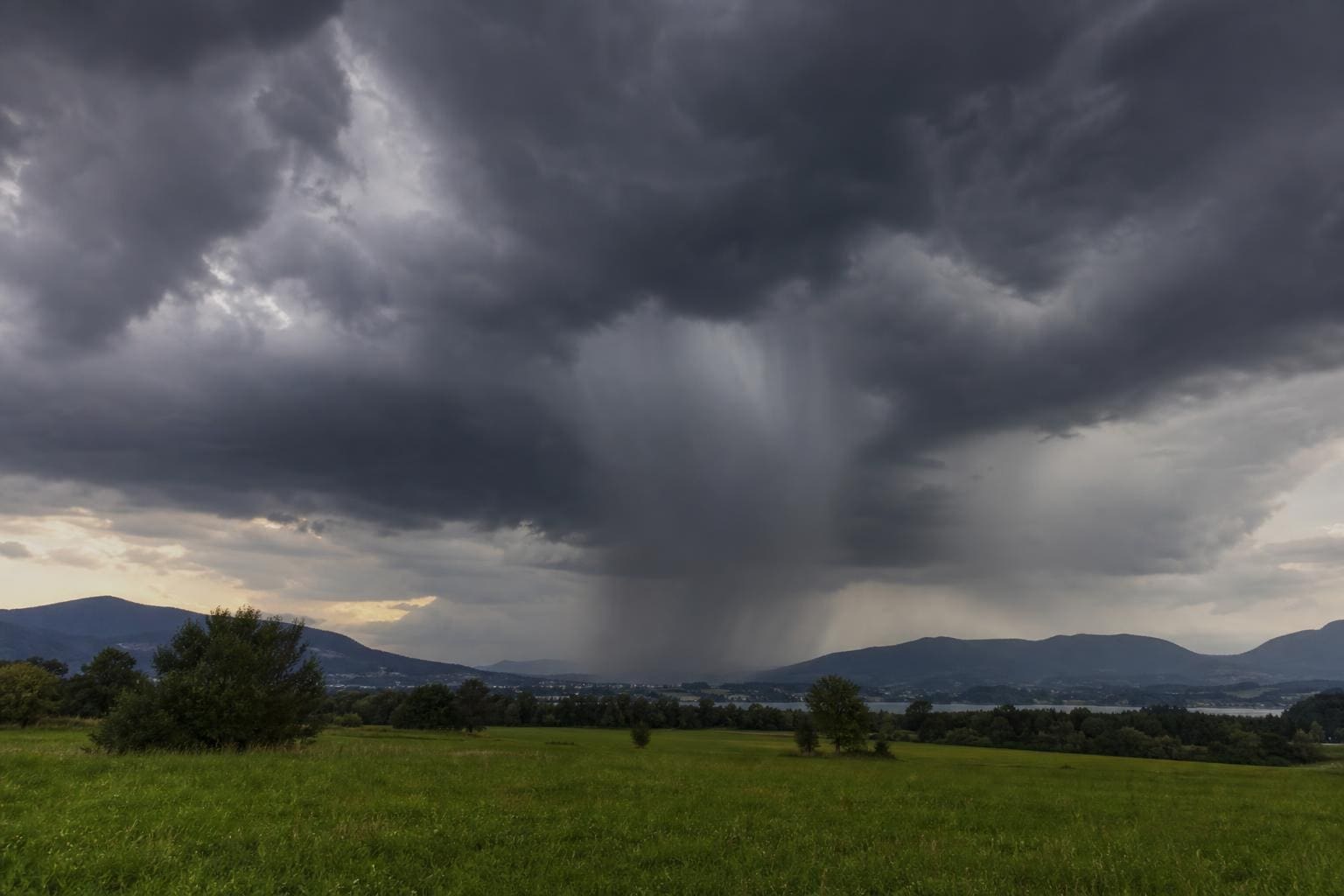 Dunkle Gewitterwolken mit Starkregen über grüner Wiese und Berglandschaft