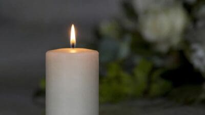 Brennende weiße Kerze im Vordergrund, unscharfe Blumen im Hintergrund