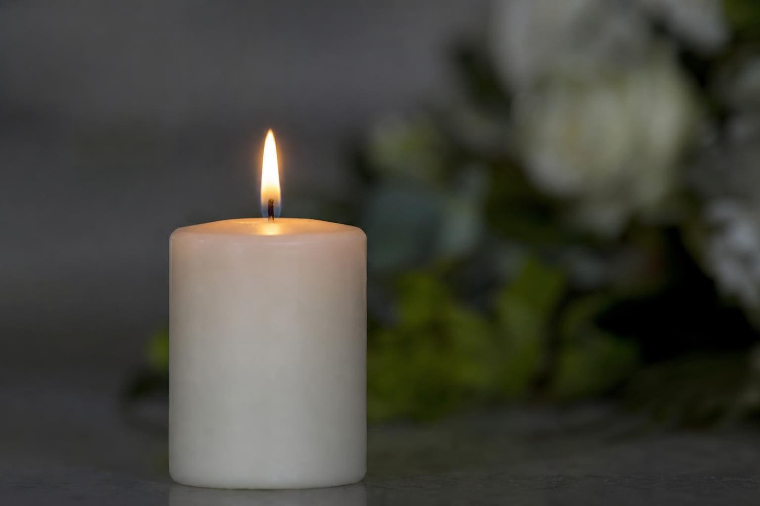Brennende weiße Kerze im Vordergrund, unscharfe Blumen im Hintergrund