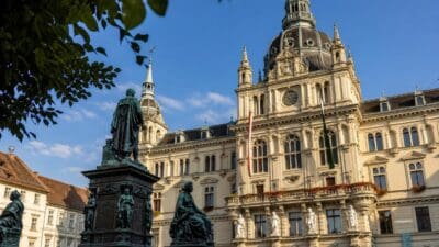 Grazer Rathaus mit Kuppel, Bronzestatuen und Fahnen unter blauem Himmel hinter Baumlaub