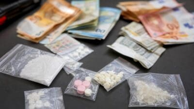 Gestapelte Euro-Banknoten und durchsichtige Tüten mit weißem Pulver sowie Tabletten auf dunkler Fläche