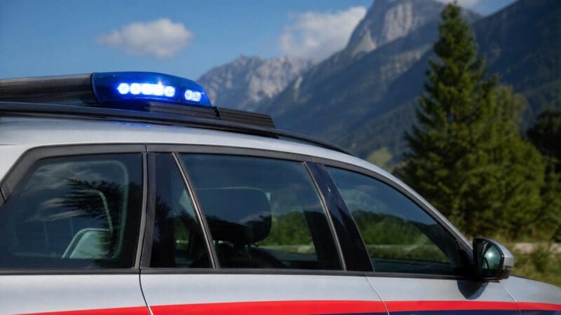 Seitenansicht eines silbernen Polizeiautos mit rot-blauen Streifen und leuchtendem Blaulicht vor Bergen