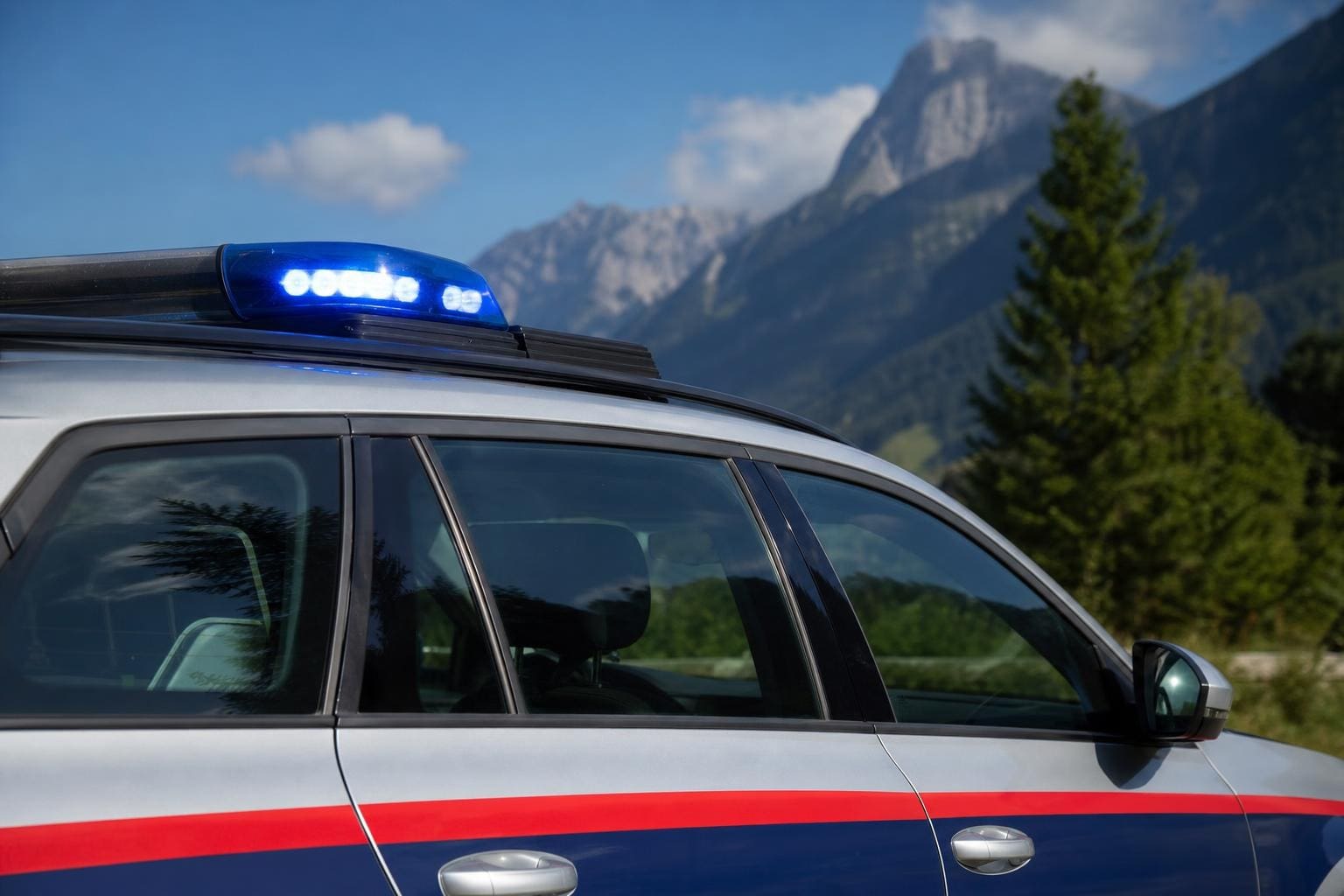 Seitenansicht eines silbernen Polizeiautos mit rot-blauen Streifen und leuchtendem Blaulicht vor Bergen