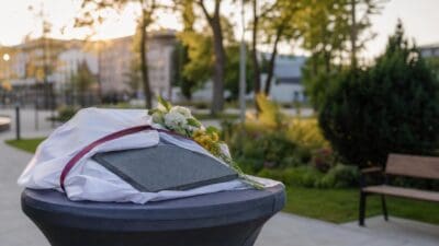 Graue Gedenktafel mit Blumen und weißem Tuch auf einem Sockel in einem Park