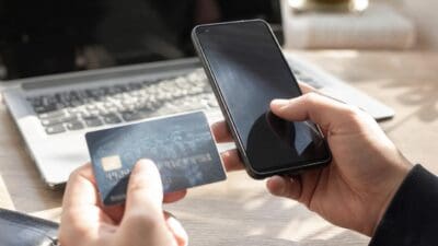 Zwei Hände halten eine Kreditkarte und ein Smartphone vor einem Laptop auf einem Holztisch.