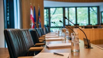 Leerer Sitzungssaal mit langem Holztisch, schwarzen Ledersesseln, Mikrofonen und Dokumenten