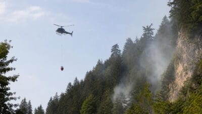 Hubschrauber mit rotem Löschwasserbehälter fliegt über rauchenden steilen Waldhang