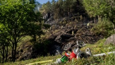 Feuerwehrschläuche und Ventile auf einer Wiese vor einem verbrannten Waldhang mit Einsatzkräften