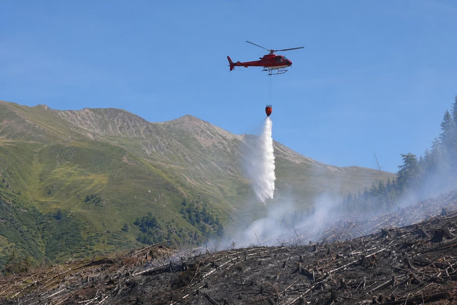 Roter Hubschrauber löscht mit Wasser aus einem Behälter einen Waldbrand in den Bergen