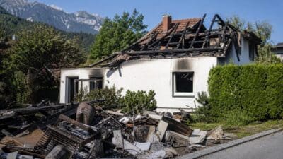 Ausgebranntes weißes Haus mit zerstörtem Dach und Schutt im Vordergrund vor Bergen