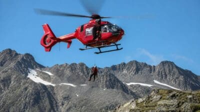 Roter Rettungshubschrauber schwebt über felsiger Berglandschaft mit einem am Seil hängenden Bergretter