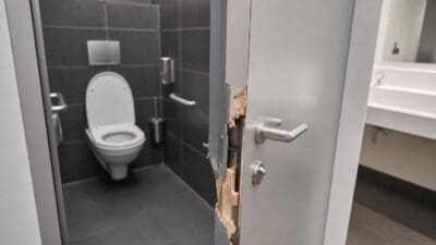 Beschädigte graue Metalltür mit zerbrochenem Rahmen vor einem weißen WC in einem Badezimmer