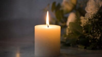 Eine brennende weiße Kerze vor einem unscharfen Blumenstrauß mit weißen Blüten