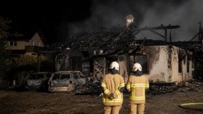 Zwei Feuerwehrleute stehen vor einem ausgebrannten Wohnhaus mit zerstörten Autos davor