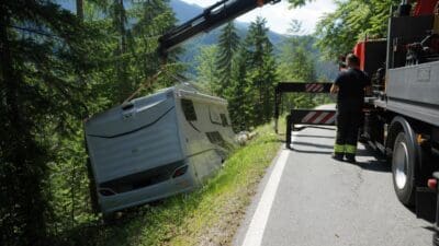 Ein weißes Wohnmobil wird von einem Kranwagen an einer Bergstraße angehoben.