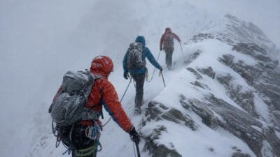 Drei Alpinisten mit Rucksäcken steigen durch Schnee und Nebel einen felsigen Grat hinauf