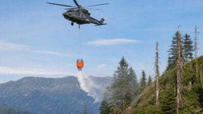 Olivgrüner Hubschrauber mit orangem Löschbehälter versprüht Wasser über bewaldetem Berggebiet
