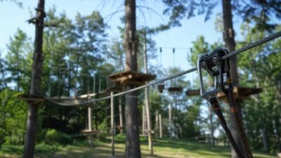 Nahaufnahme eines silbernen Karabinerhakens mit schwarzen Gurten an einem Stahlseil in einem Hochseilgarten.