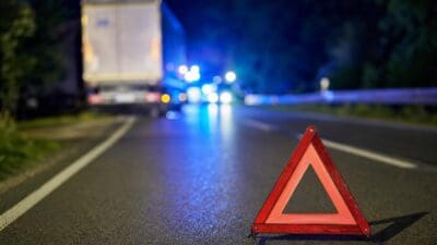 Rotes Warndreieck auf nasser Asphaltstraße bei Nacht mit Lkw und Blaulicht im Hintergrund