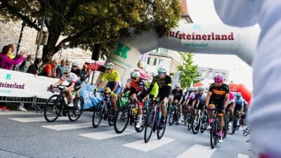 Radrennfahrer in bunten Trikots fahren unter einem großen Aufblasbogen in Kufstein hindurch