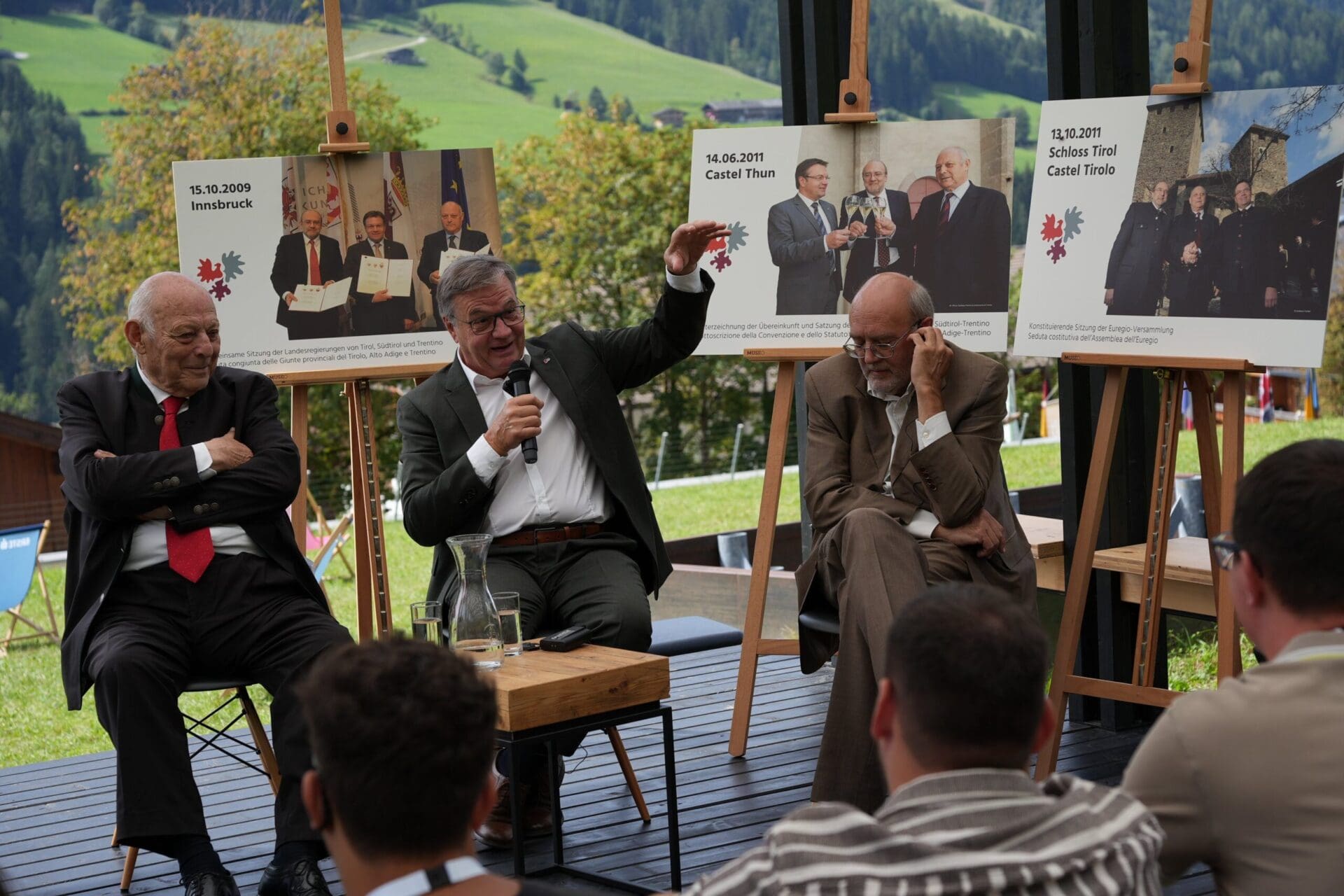 Drei ältere Männer in Anzügen auf einer Bühne, einer spricht ins Mikrofon vor historischen Fotos.