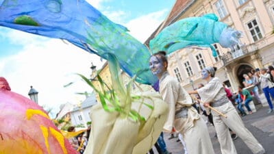 Zwei Performerinnen mit blauer Gesichtsfarbe führen eine Performance mit langen bunten Stoffen in Klagenfurt auf.