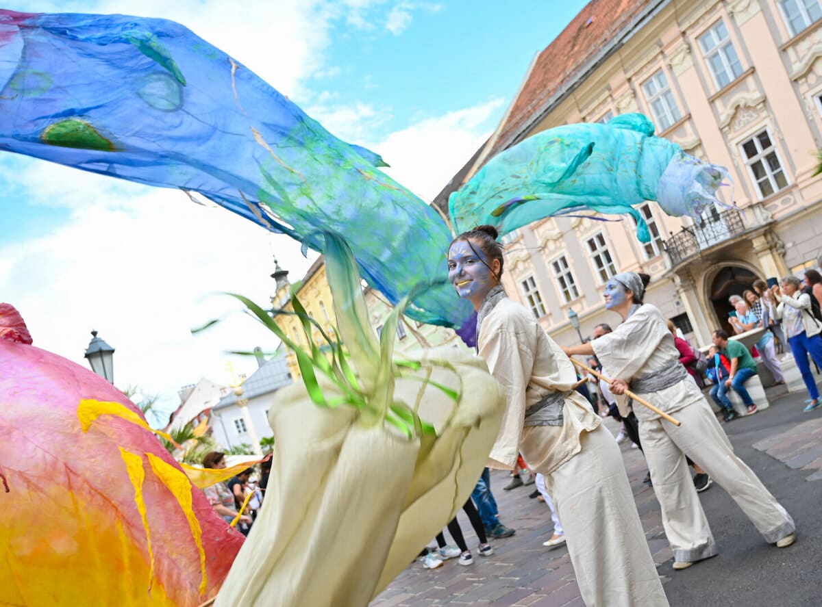 Zwei Performerinnen mit blauer Gesichtsfarbe führen eine Performance mit langen bunten Stoffen in Klagenfurt auf.