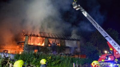 Brennendes Wirtschaftsgebäude bei Nacht mit ausgefahrener Feuerwehrdrehleiter und Einsatzkräften im Vordergrund