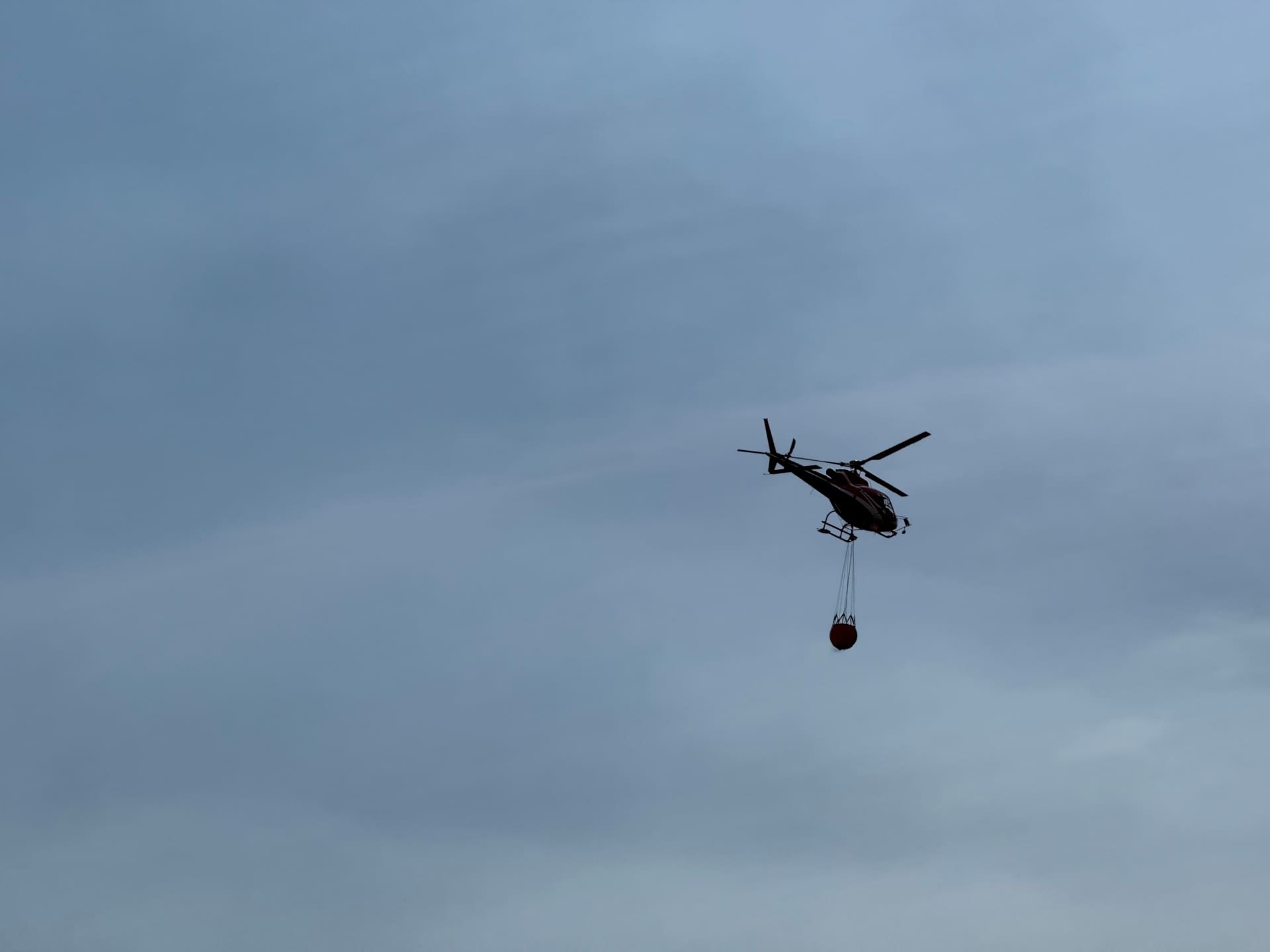 Ein Hubschrauber mit einem roten Behälter fliegt vor einem bewölkten Himmel.