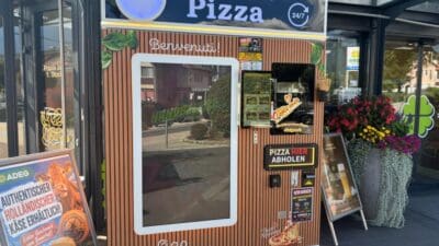 Ein braun gestreifter Pizzaautomat mit Bildschirm und Werbeaufklebern steht vor einem Supermarkteingang.