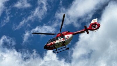 Roter und weißer Notarzt-Hubschrauber der ARA fliegt vor blauem Himmel mit Wolken.