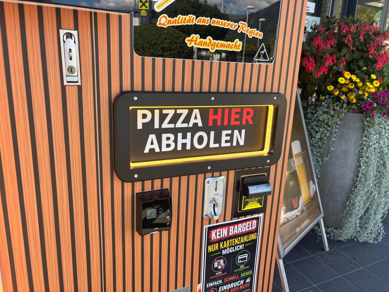 Vorderansicht eines orange-braun gestreiften Pizzaautomaten mit beleuchtetem Schild Pizza hier abholen und Zahlungsterminals.