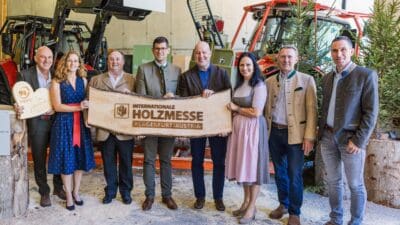 Sieben Personen halten großes Holzschild der Internationalen Holzmesse vor Forstmaschinen in Halle.